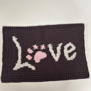 May include: A dark brown crocheted rectangular item with the word "Love" in white and a pink paw print. The item is likely a decorative piece or a small mat, showcasing a handmade craft.