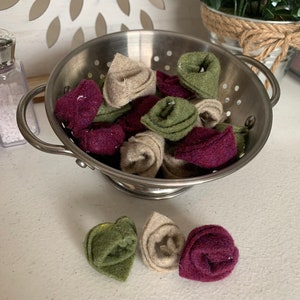 May include: A metal colander filled with felt fabric flowers in shades of burgundy, green, and beige. The flowers are arranged in a random pattern.
