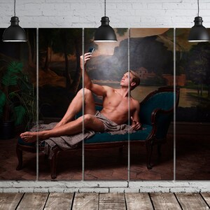 May include: A man in a reclining pose on a chaise lounge, taking a selfie with a smartphone. He is wearing a white cloth draped around his waist. The background is a painted mural of a landscape with trees and a river.