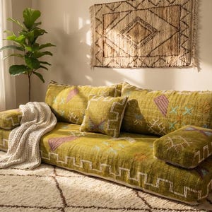 May include: A low-profile olive green floor sofa featuring a textured fabric with white geometric patterns and embroidered accents. The sofa includes two rectangular bolster pillows, a square throw pillow, and a cream-colored knit throw blanket.