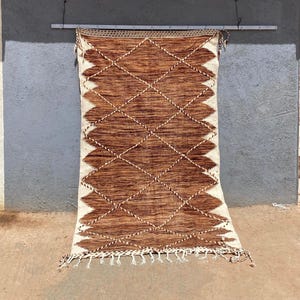 May include: A brown and cream-colored woven rug with a diamond pattern. The rug has a geometric design with diamond shapes and a zigzag border. The rug is hanging against a gray wall and has tassels along the bottom edge.