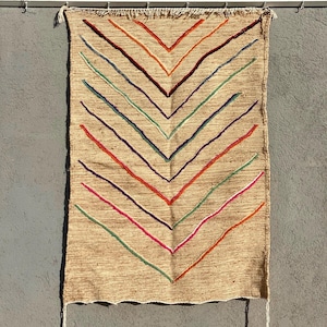 Könnte beinhalten: Ein beigefarbener gewebter Wandbehang mit Zickzackmuster. Das Design zeigt farbige Linien in Rot, Orange, Lila, Blau, Grün und Rosa. Der obere Rand hat eine Fransenverzierung, und das Stück wird an einer Metallstange aufgehängt.