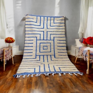 Può includere: Un grande tappeto rettangolare con un design geometrico. Il tappeto presenta uno sfondo color crema con un audace motivo blu di quadrati e linee. Il bordo e le nappe sono blu. Il tappeto è esposto su un pavimento in legno.