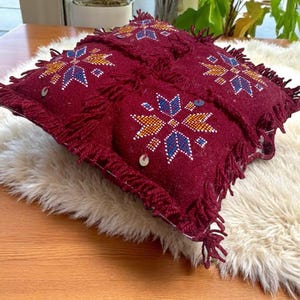 Può includere: Un cuscino decorativo bordeaux con un design geometrico blu, giallo e bianco. Il cuscino ha un bordo con frange ed è impreziosito da paillettes. È appoggiato su una superficie bianca e soffice.