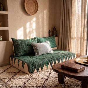 May include: A low-profile floor sofa featuring a dark green textured cover with a repeating geometric pattern and a cream-colored base. The sofa is accented with two large green rectangular pillows and one smaller, smooth light gray pillow.