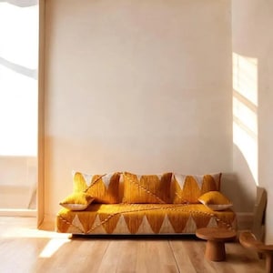 Può includere: Un divano giallo senape con un motivo geometrico di triangoli bianchi. Il divano ha cuscini abbinati e un tavolino in legno. La stanza ha pareti chiare e un pavimento in legno.