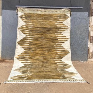 Pode incluir: Um tapete tecido com um design geométrico. O tapete apresenta um padrão de diamante central em tons de castanho e dourado, ladeado por ziguezagues brancos. O tapete está pendurado contra uma parede cinzenta.