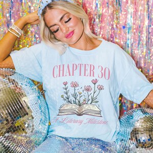 May include: A light blue t-shirt with a pink floral design and the text "CHAPTER 30 A Literary Milestone".