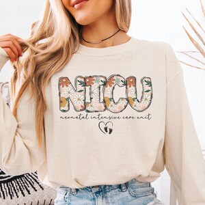 May include: A cream-colored long-sleeved shirt with the words "NICU" in floral letters and the text "neonatal intensive care unit" below. The shirt also features a small heart with footprints.