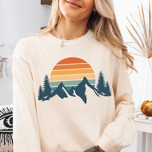 May include: A cream colored long-sleeved sweatshirt with a retro sunset and mountain graphic. The graphic features a blue silhouette of mountains with a sunset behind them in shades of orange, yellow, and red.