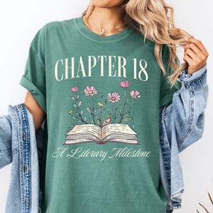 May include: A green t-shirt with the text "CHAPTER 18" above an open book illustration with flowers. The text "A Literary Milestone" is below the book. The shirt is a solid green color.
