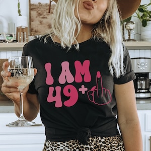 May include: A black t-shirt with the text "I AM 49+" in pink and a pink middle finger graphic.