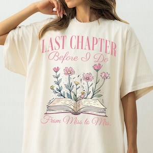 May include: Cream-colored t-shirt with the text "LAST CHAPTER Before I Do From Miss to Mrs." in pink. The shirt features a floral design with pink flowers and an open book. The shirt has short sleeves and a relaxed fit.