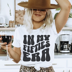 May include: White t-shirt with black text that reads "In My Fifties Era" in a retro font. The person wearing the shirt is holding a glass of red wine and wearing a brown hat.