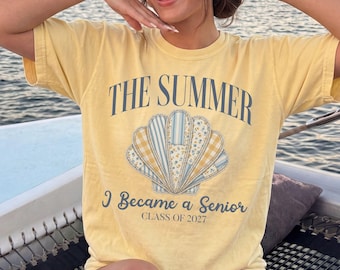 May include: A pale yellow short-sleeve t-shirt featuring a graphic of a seashell with blue, white, and yellow patterned segments. The text reads THE SUMMER I Became a Senior CLASS OF 2027 in dark blue lettering across the front of the garment.