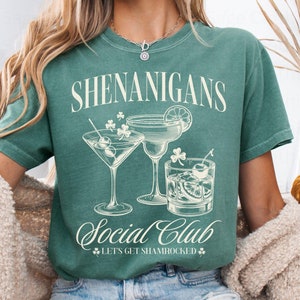 May include: A teal green t-shirt with the text "Shenanigans Social Club" and a graphic of three cocktail glasses. The text below the glasses reads "Let's Get Shamrocked".
