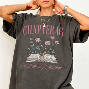 May include: A dark gray oversized t-shirt with a graphic print of a book with flowers growing out of it. The text on the t-shirt reads 'Chapter 16 A Literary Milestone'.
