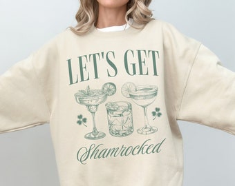 Puede incluir: Una sudadera beige con el texto "Let's Get Shamrocked" en letras verdes. El gráfico presenta tres ilustraciones verdes de cócteles.