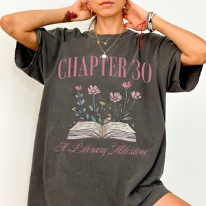 May include: A dark gray oversized t-shirt with a graphic print of an open book with flowers and the text "CHAPTER 30 A Literary Milestone".