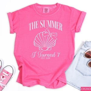 K&ouml;nnte beinhalten: Ein leuchtend pinkfarbenes T-Shirt mit dem wei&szlig;en Aufdruck "THE SUMMER I TURNED 7 EST 2019" und einer Muschel-Grafik. Dazu geh&ouml;ren hellblaue Jeansshorts, eine rosa Sonnenbrille und rosa Turnschuhe.