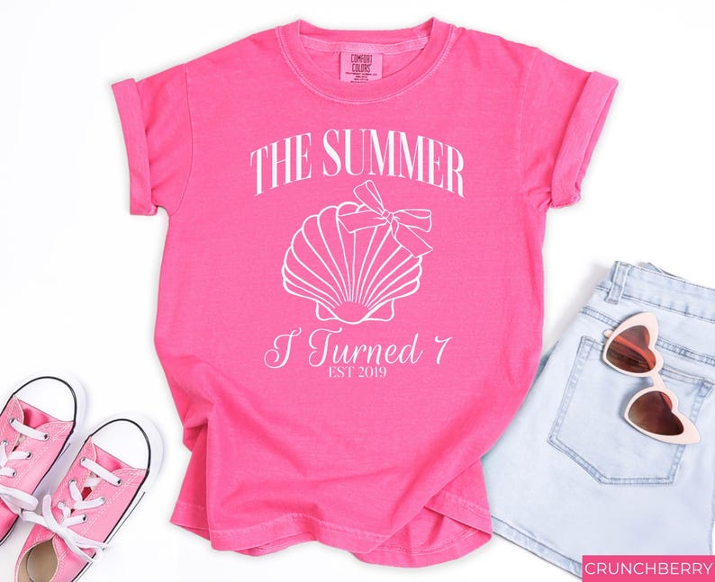 K&ouml;nnte beinhalten: Ein leuchtend pinkfarbenes T-Shirt mit dem wei&szlig;en Aufdruck "THE SUMMER I TURNED 7 EST 2019" und einer Muschel-Grafik. Dazu geh&ouml;ren hellblaue Jeansshorts, eine rosa Sonnenbrille und rosa Turnschuhe.