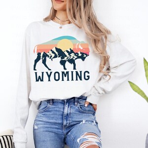 May include: White long-sleeve sweatshirt with a retro-style graphic of a buffalo silhouette in front of mountains and a sun. The text "WYOMING" is printed below the graphic.