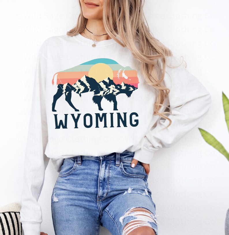May include: White long-sleeve sweatshirt with a retro-style graphic of a buffalo silhouette in front of mountains and a sun. The text "WYOMING" is printed below the graphic.