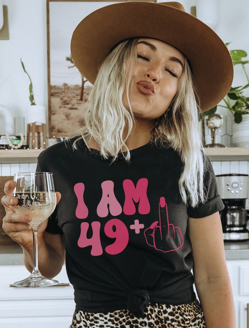 May include: A black t-shirt with the text "I AM 49+" in pink and a pink middle finger graphic.