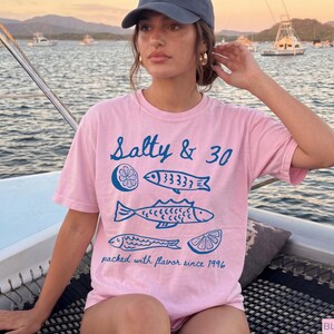 May include: A pale pink t-shirt with the words "Salty & 30" and blue fish and lemon slice illustrations. The text "packed with flavour since 1996" is also printed on the shirt. The person is wearing a blue cap and gold earrings.
