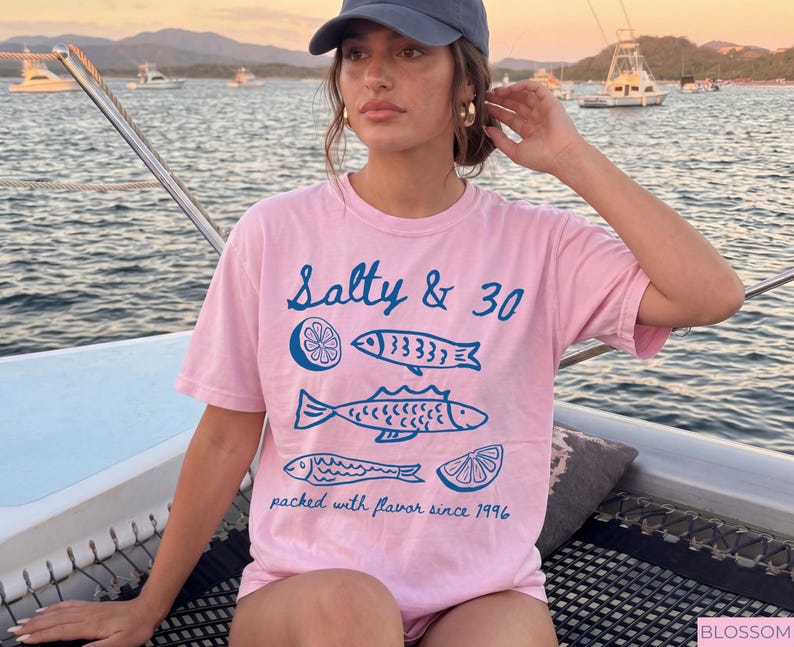 May include: A pale pink t-shirt with the words "Salty & 30" and blue fish and lemon slice illustrations. The text "packed with flavour since 1996" is also printed on the shirt. The person is wearing a blue cap and gold earrings.