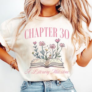 May include: A cream colored t-shirt with the text "CHAPTER 30 A Literary Milestone" in pink lettering. The design features a book with pink flowers growing from it.