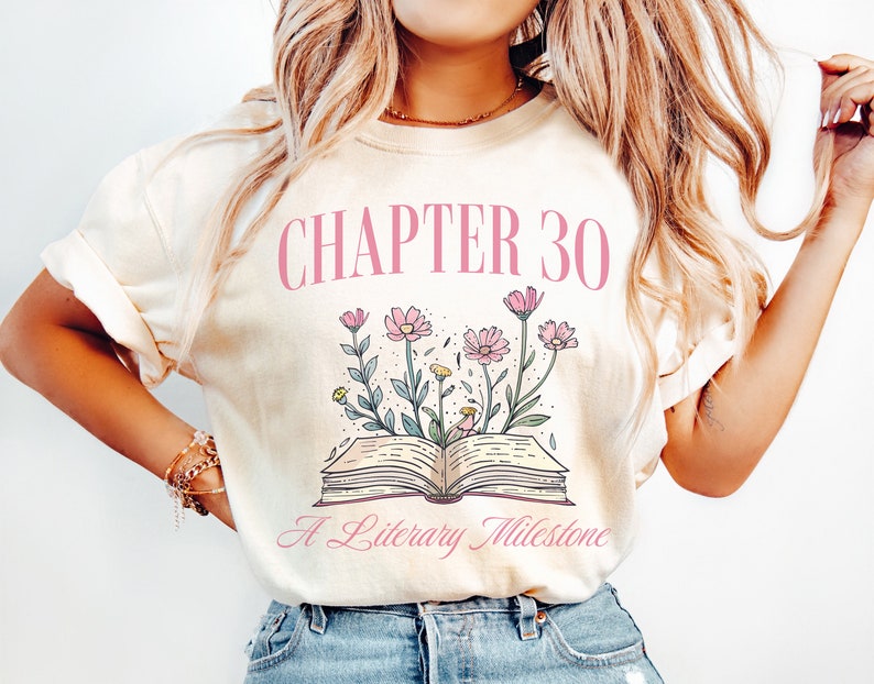 May include: A cream colored t-shirt with the text "CHAPTER 30 A Literary Milestone" in pink lettering. The design features a book with pink flowers growing from it.