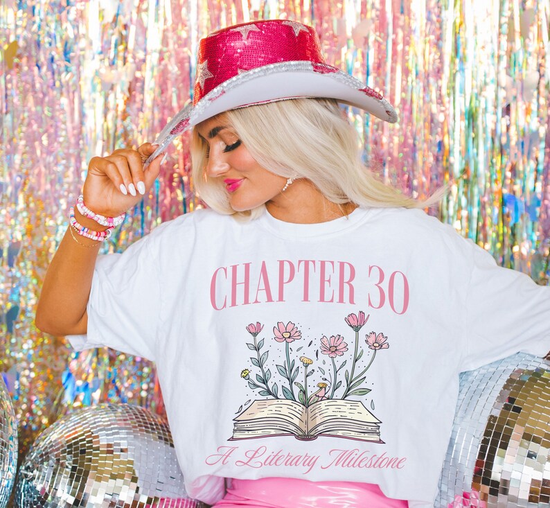 May include: White t-shirt with pink text that reads "CHAPTER 30 A Literary Milestone". The shirt features a pink open book with flowers growing from it.
