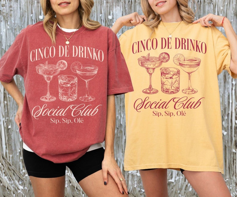 May include: Two women wearing oversized t-shirts with the text "Cinco de Drinko Social Club Sip, Sip, Ol&eacute;" and a graphic of cocktail glasses. The shirt on the left is red and the shirt on the right is yellow.