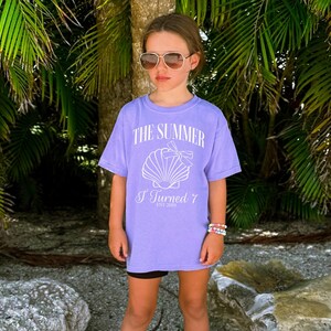 K&ouml;nnte beinhalten: Ein violettes T-Shirt mit dem Text "THE SUMMER I Turned 7 EST 2019" und einer Muschel-Grafik. Das Shirt wird mit schwarzen Shorts und Sonnenbrille getragen. Der Hintergrund zeigt Palmen und eine Strandkulisse.