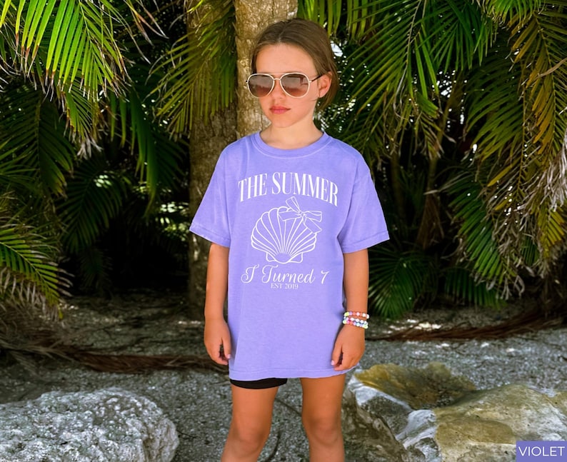 K&ouml;nnte beinhalten: Ein violettes T-Shirt mit dem Text "THE SUMMER I Turned 7 EST 2019" und einer Muschel-Grafik. Das Shirt wird mit schwarzen Shorts und Sonnenbrille getragen. Der Hintergrund zeigt Palmen und eine Strandkulisse.