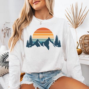May include: White long-sleeve sweatshirt with a retro sunset and mountain graphic. The graphic features a blue, orange, and yellow sunset with a silhouette of mountains and trees.
