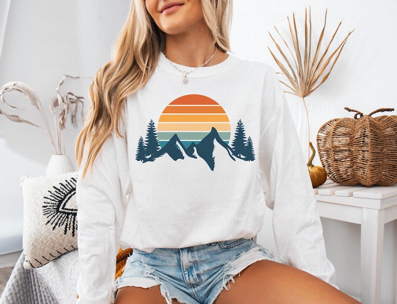 May include: White long-sleeve sweatshirt with a retro sunset and mountain graphic. The graphic features a blue, orange, and yellow sunset with a silhouette of mountains and trees.