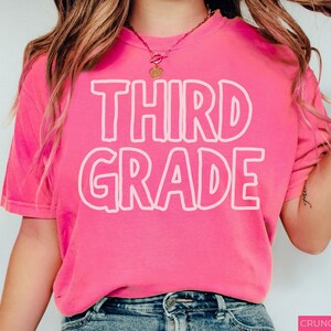 Pu&ograve; includere: Una t-shirt a maniche corte di colore rosa acceso con la scritta THIRD GRADE stampata in un carattere bianco con contorno spesso sul petto. Il capo presenta un girocollo ed &egrave; abbinato a jeans blu, creando un aspetto casual e vivace.