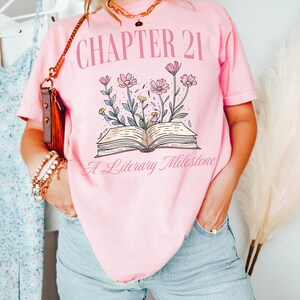 May include: Pink t-shirt with the text "Chapter 21 A Literary Milestone" and a graphic of an open book with flowers growing from it.