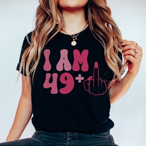 May include: Black t-shirt with a pink and white graphic that says "I AM 49+" with a middle finger emoji.