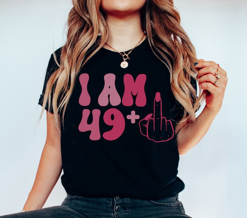 May include: Black t-shirt with a pink and white graphic that says "I AM 49+" with a middle finger emoji.