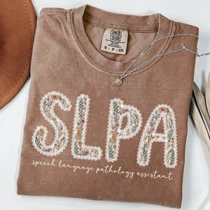 May include: A brown Comfort Colors t-shirt with the letters "SLPA" embroidered with floral designs. The text "speech language pathology assistant" is below the letters. A silver necklace with a pendant is on the shirt.
