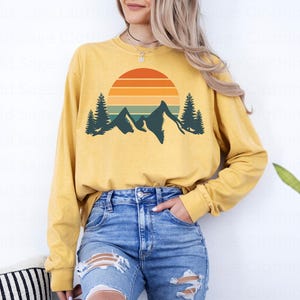 May include: A yellow long-sleeved shirt with a retro sunset and mountain graphic. The graphic features a sun with orange, red, and yellow stripes, a mountain range, and two pine trees.