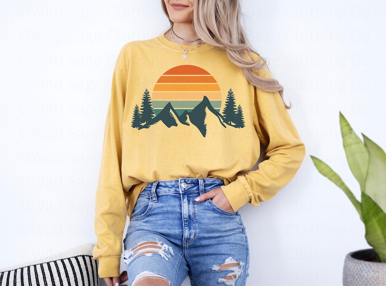May include: A yellow long-sleeved shirt with a retro sunset and mountain graphic. The graphic features a sun with orange, red, and yellow stripes, a mountain range, and two pine trees.