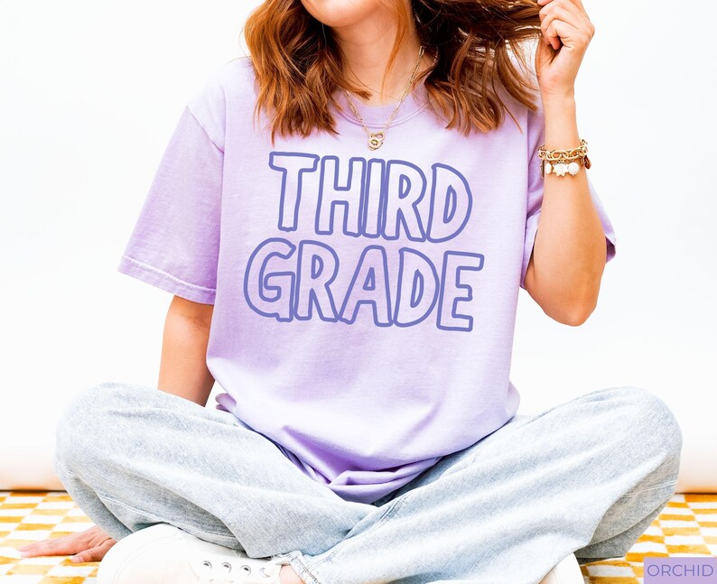 Pu&ograve; includere: Una t-shirt a maniche corte color viola chiaro con la scritta THIRD GRADE in un carattere audace con contorno viola scuro. La maglia &egrave; indossata con jeans azzurri, una collana a catena dorata e un braccialetto color oro con perline bianche.