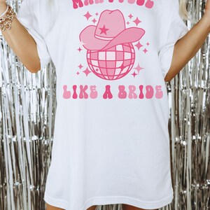 K&ouml;nnte beinhalten: Wei&szlig;es Oversize-T-Shirt mit einem rosafarbenen Grafikdesign. Die Grafik zeigt eine Discokugel mit einem rosafarbenen Cowboyhut darauf. Der Text "Man I Feel Like A Bride" ist in rosafarbenen Buchstaben geschrieben.