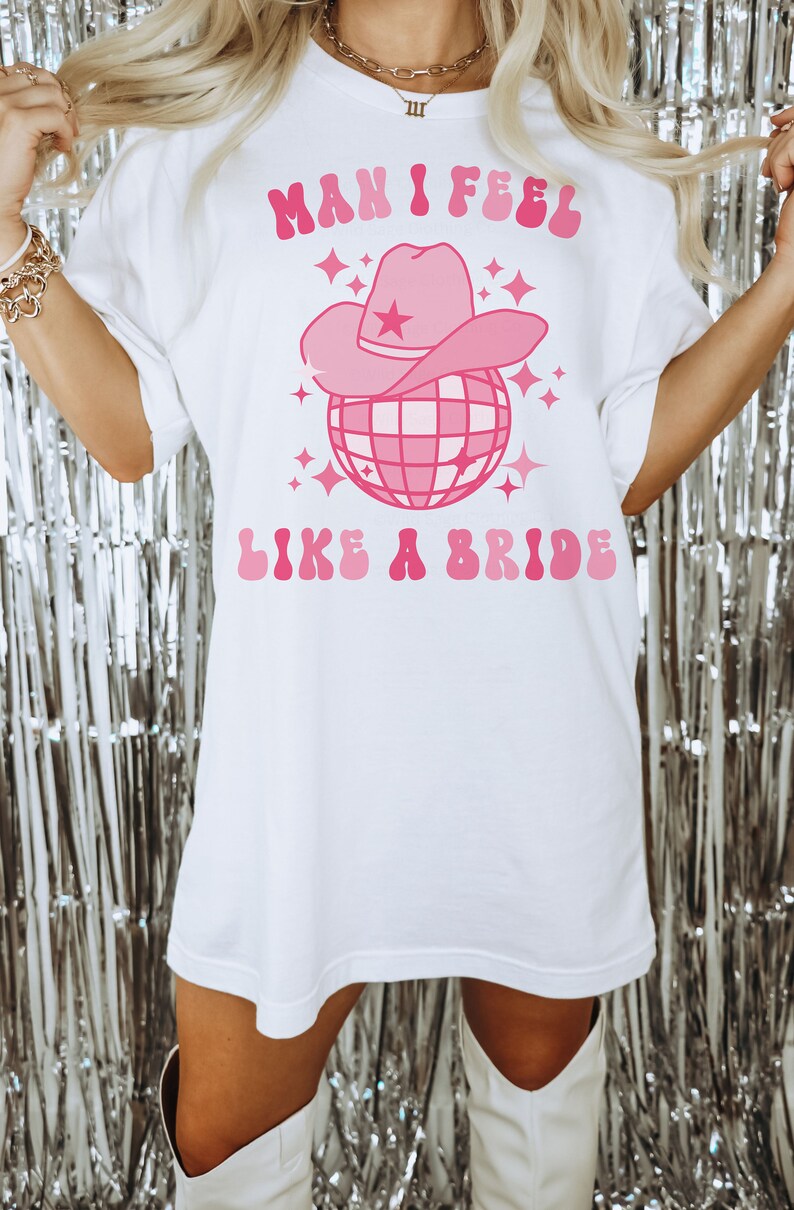 K&ouml;nnte beinhalten: Wei&szlig;es Oversize-T-Shirt mit einem rosafarbenen Grafikdesign. Die Grafik zeigt eine Discokugel mit einem rosafarbenen Cowboyhut darauf. Der Text "Man I Feel Like A Bride" ist in rosafarbenen Buchstaben geschrieben.