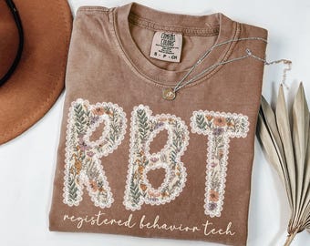 May include: A brown Comfort Colors t-shirt with the letters "RBT" in a floral design. The words "registered behavior tech" are printed below the letters. A silver necklace with a pendant is draped across the shirt.