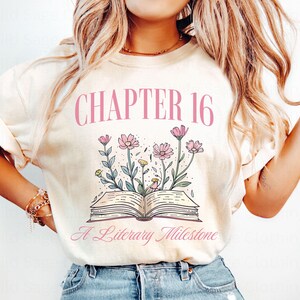 Puede incluir: Una camiseta de color crema con el texto "CHAPTER 16 A Literary Milestone" en rosa. La camiseta presenta un dise&ntilde;o floral rosa con un libro en el centro.
